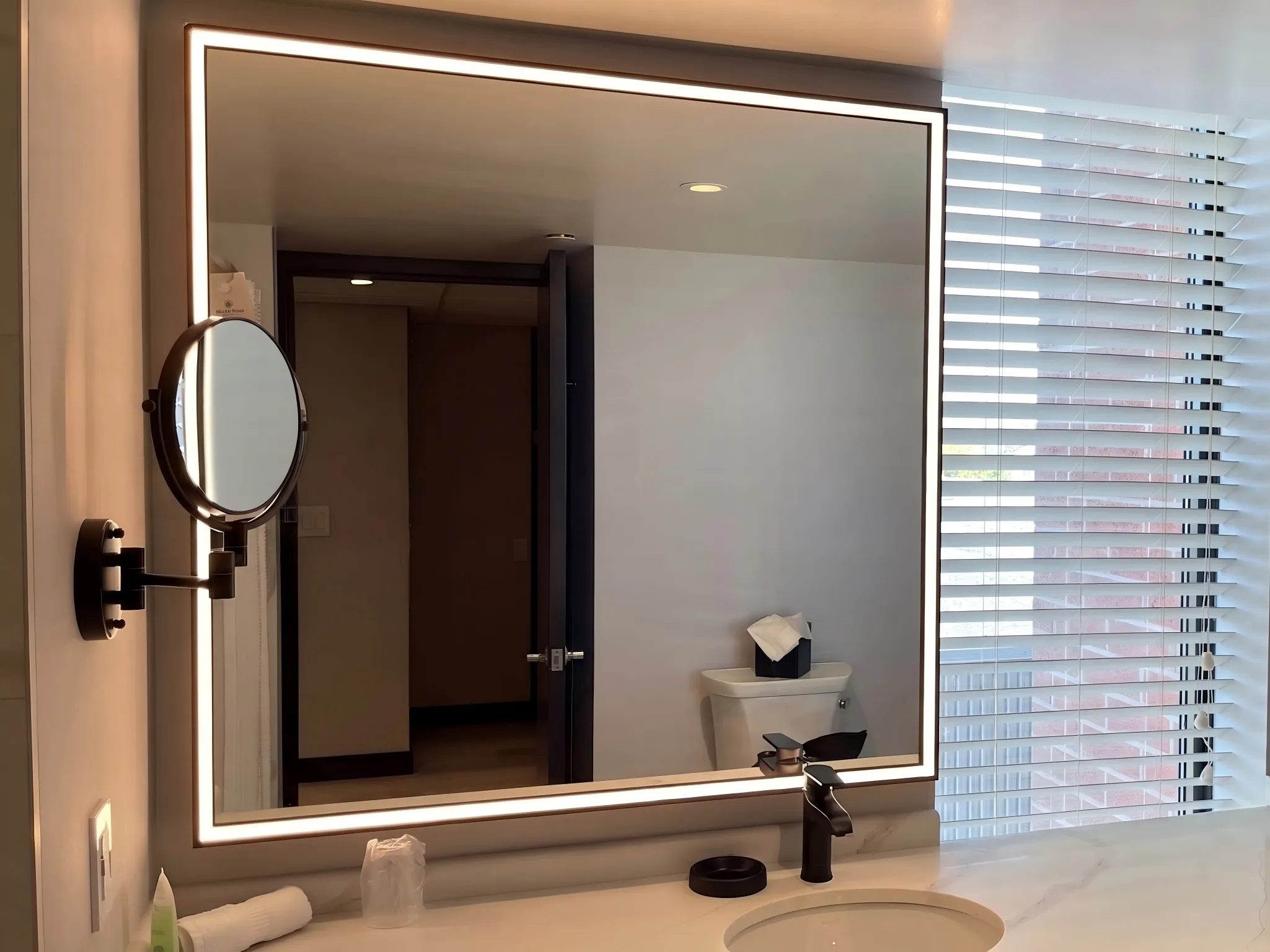 A professionally installed custom rectangular LED backlit mirror in a modern California hotel guest bathroom, featuring an unobstructed reflection of the room showing blinds and a concrete pillar. The mirror is ETL/UL certified for commercial hospitality use.